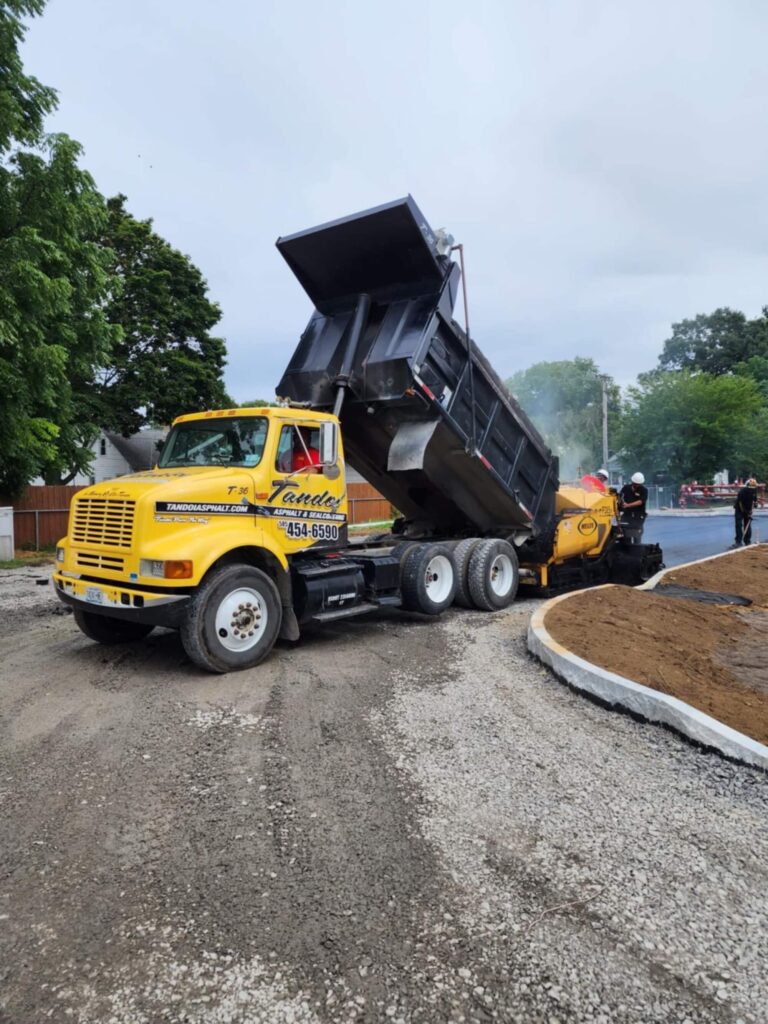 A yellow dump truck is unloading asphalt into a paving machine on a road construction site, with fresh pavement and gravel visible. Trees and workers are in the background.