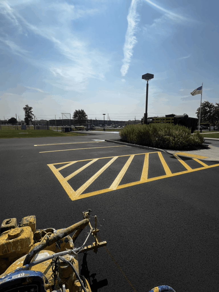 A freshly painted yellow-striped parking area on black asphalt under a blue sky with wispy clouds, an American flag, and some greenery in the background. A paint striping machine is visible in the foreground.