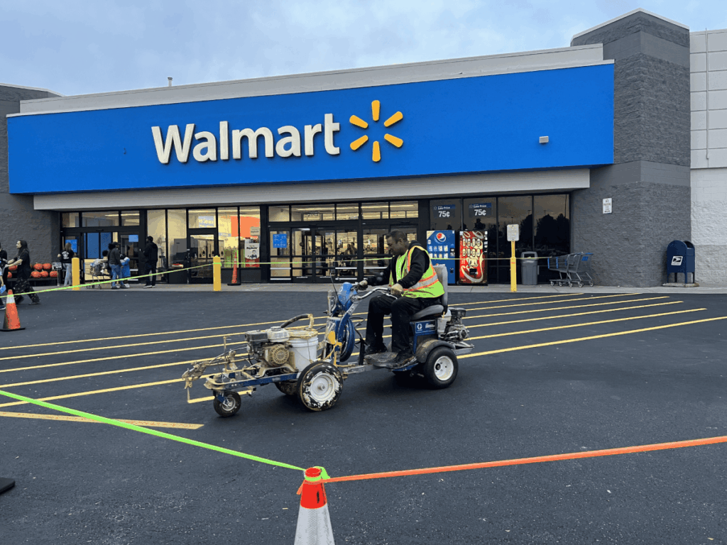 A worker on a line striping machine paints new parking lines in front of a Walmart store. Orange cones and caution tape block off the area. People and shopping carts are near the entrance.