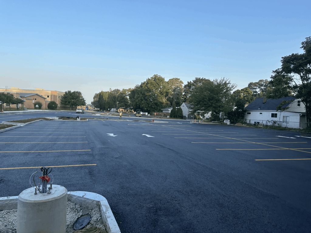 A freshly paved, empty parking lot with newly painted yellow lines and white arrows, surrounded by trees, a few houses, and a brick building under a clear blue sky.