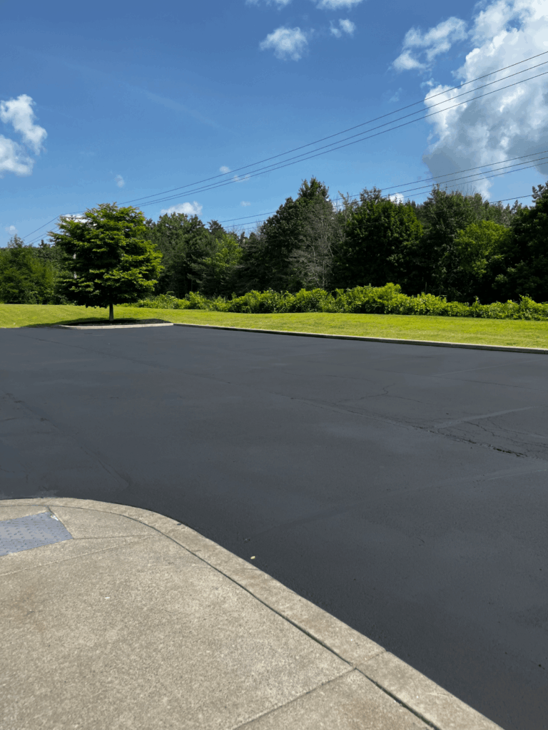 A freshly paved, empty parking lot under a bright blue sky with scattered clouds. There is a grassy area with a lone tree and green shrubs in the background, alongside power lines.