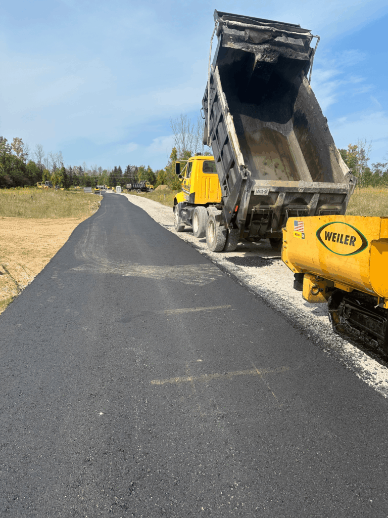A yellow dump truck unloads asphalt onto a rural road under construction, while a yellow paving machine spreads the asphalt. The road stretches into the distance with fields and trees on both sides.