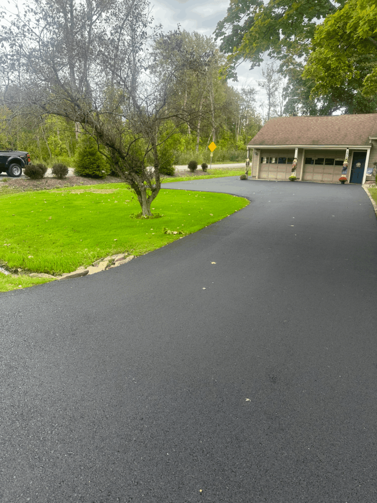Freshly paved black asphalt driveway curves toward a three-car garage attached to a house. Green lawn and trees surround the driveway; a parked truck and road are visible in the background.