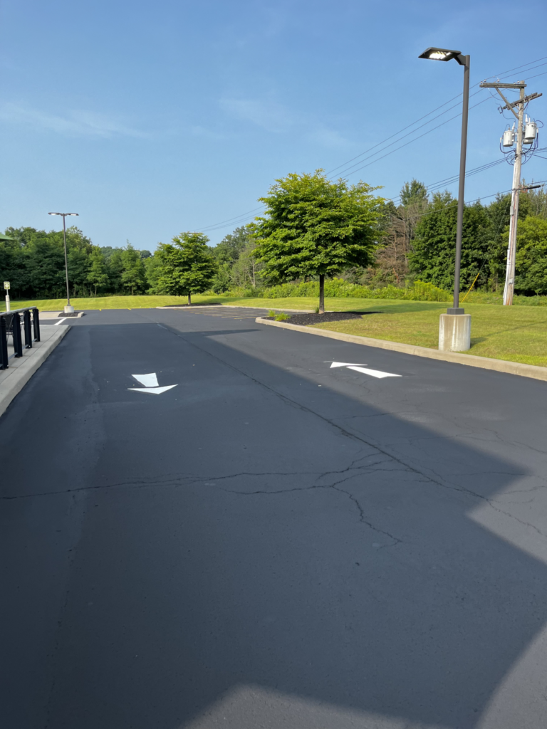 A freshly paved parking lot with two white directional arrows, one pointing straight and one pointing right. Green trees, grass, and a blue sky are visible in the background, with streetlights and utility poles on the right.