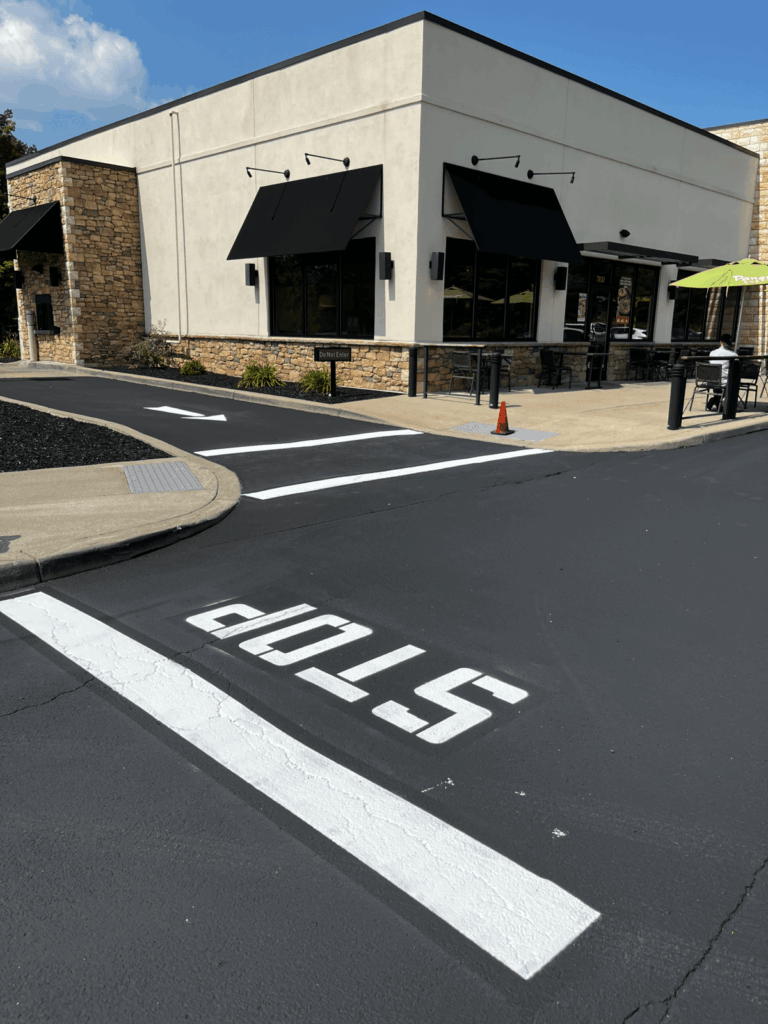 A modern commercial building with black awnings sits beside a freshly paved drive-thru lane. The word STOP is painted on the pavement near a white stop line. An orange cone stands near the sidewalk.