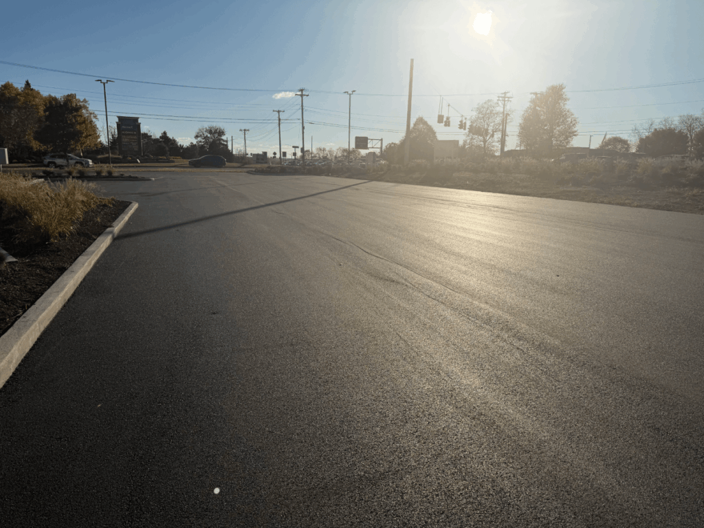 Freshly paved empty parking lot in the early morning or late afternoon sunlight, with shadows, a curb, some grass to the left, and trees, power lines, and traffic lights visible in the background.