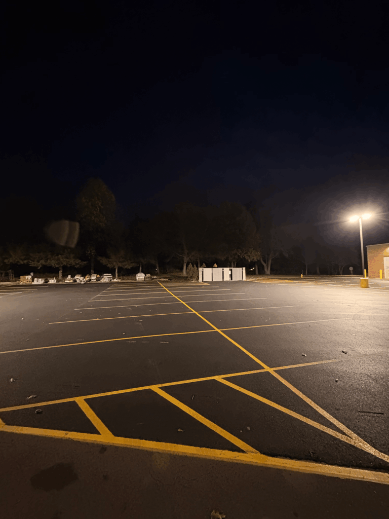 An empty parking lot at night is illuminated by a single streetlamp on the right. Yellow parking lines are visible, with trees and a white utility shed in the background under a dark sky.