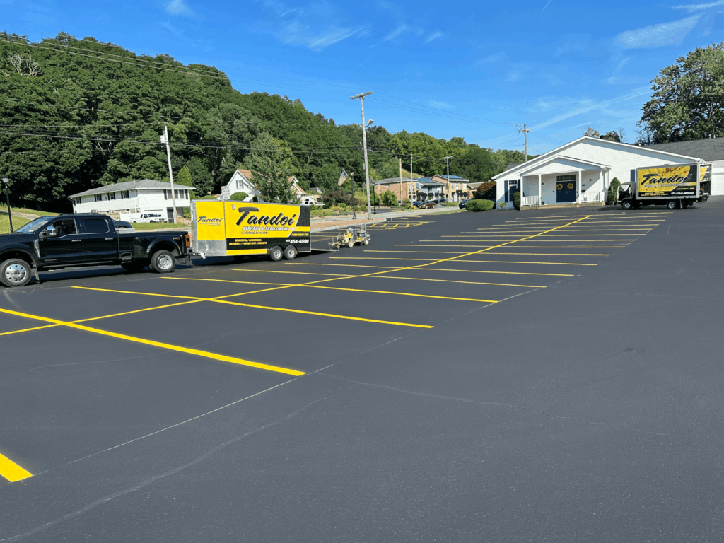 A freshly paved and striped parking lot with bright yellow lines. Two vehicles with yellow “Tandol” company trailers are parked nearby on a sunny day, with trees and buildings in the background.