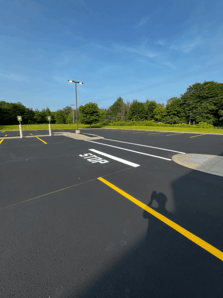 An empty parking lot with fresh black pavement, yellow parking lines, a white STOP marking, two parking signs, a streetlight, and green trees under a clear blue sky. A persons shadow is visible in the foreground.